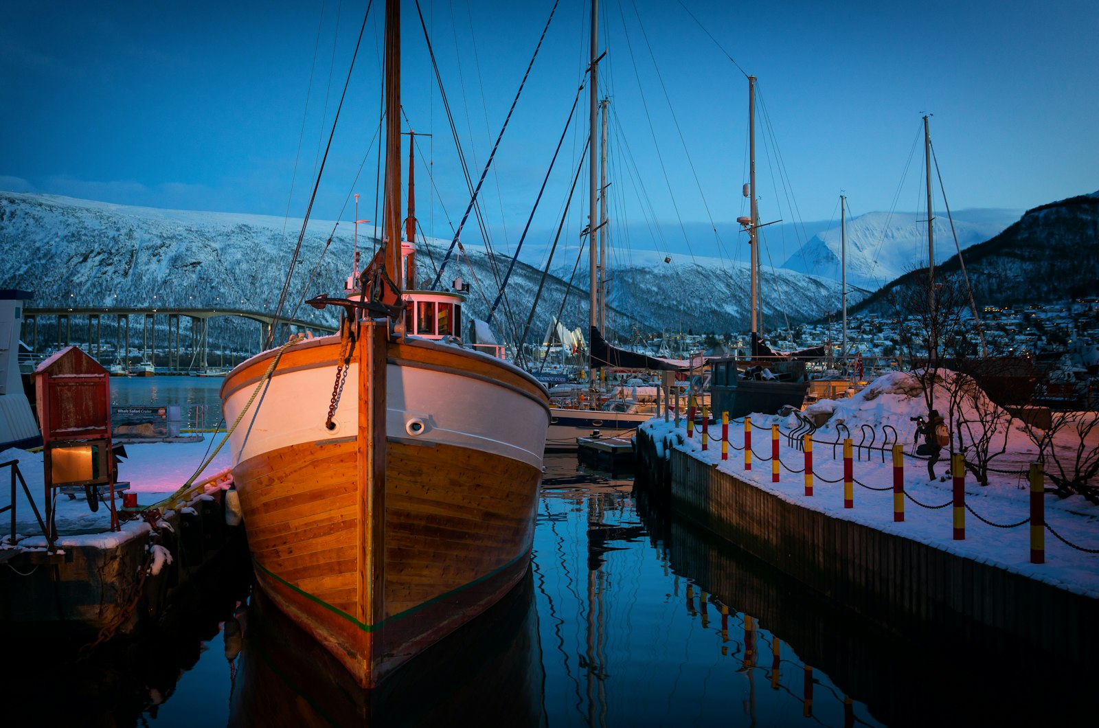 Trebåt fortøyd i snødekt havn i Tromsø med fjell og bro i bakgrunnen